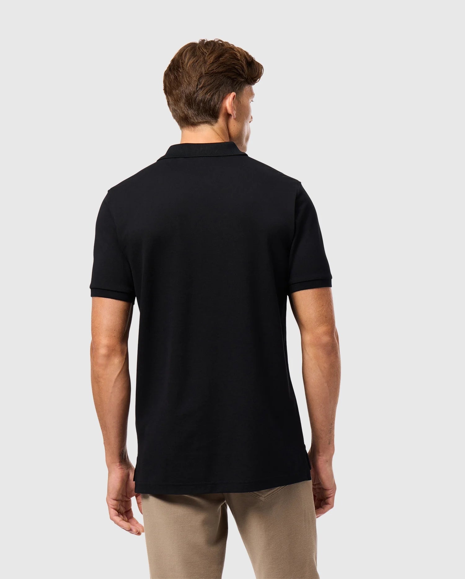 A man with short brown hair is standing with his back to the camera, donning a plain black Psycho Bunny MENS JAMES PREMIUM PIQUE POLO SHIRT - B6K945D200 made of durable Pima cotton and beige pants. The background is plain and light grey.