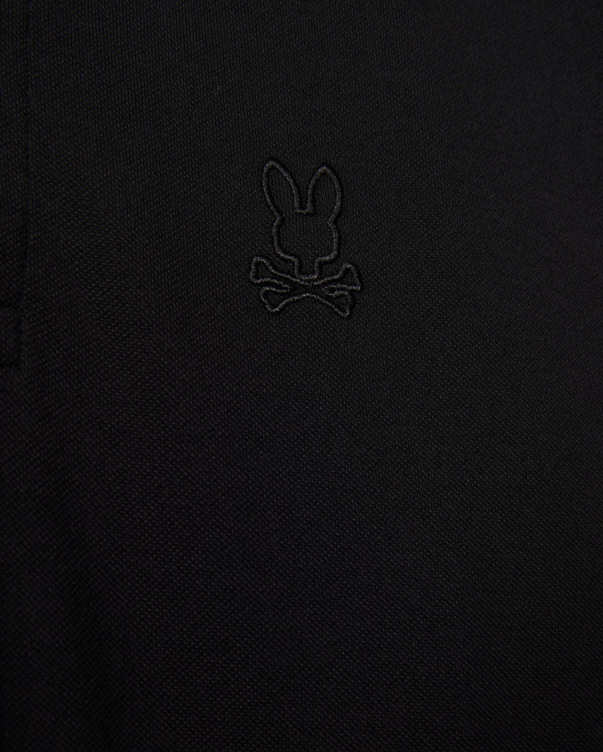 Close-up of the Psycho Bunny MENS JAMES PREMIUM PIQUE POLO SHIRT - B6K945D200 in black, showcasing an embroidered logo depicting a rabbit's head with crossbones underneath. The all-black logo blends subtly with the fabric, adding a refined touch. Made from premium Pima cotton, this durable shirt offers both style and comfort.