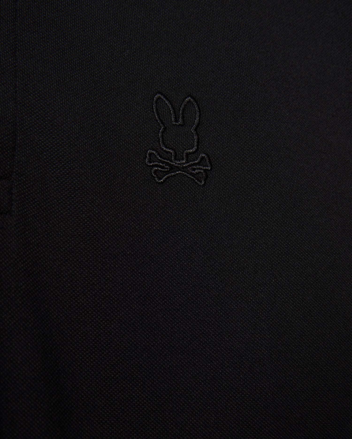 Close-up of the Psycho Bunny MENS JAMES PREMIUM PIQUE POLO SHIRT - B6K945D200 in black, showcasing an embroidered logo depicting a rabbit's head with crossbones underneath. The all-black logo blends subtly with the fabric, adding a refined touch. Made from premium Pima cotton, this durable shirt offers both style and comfort.