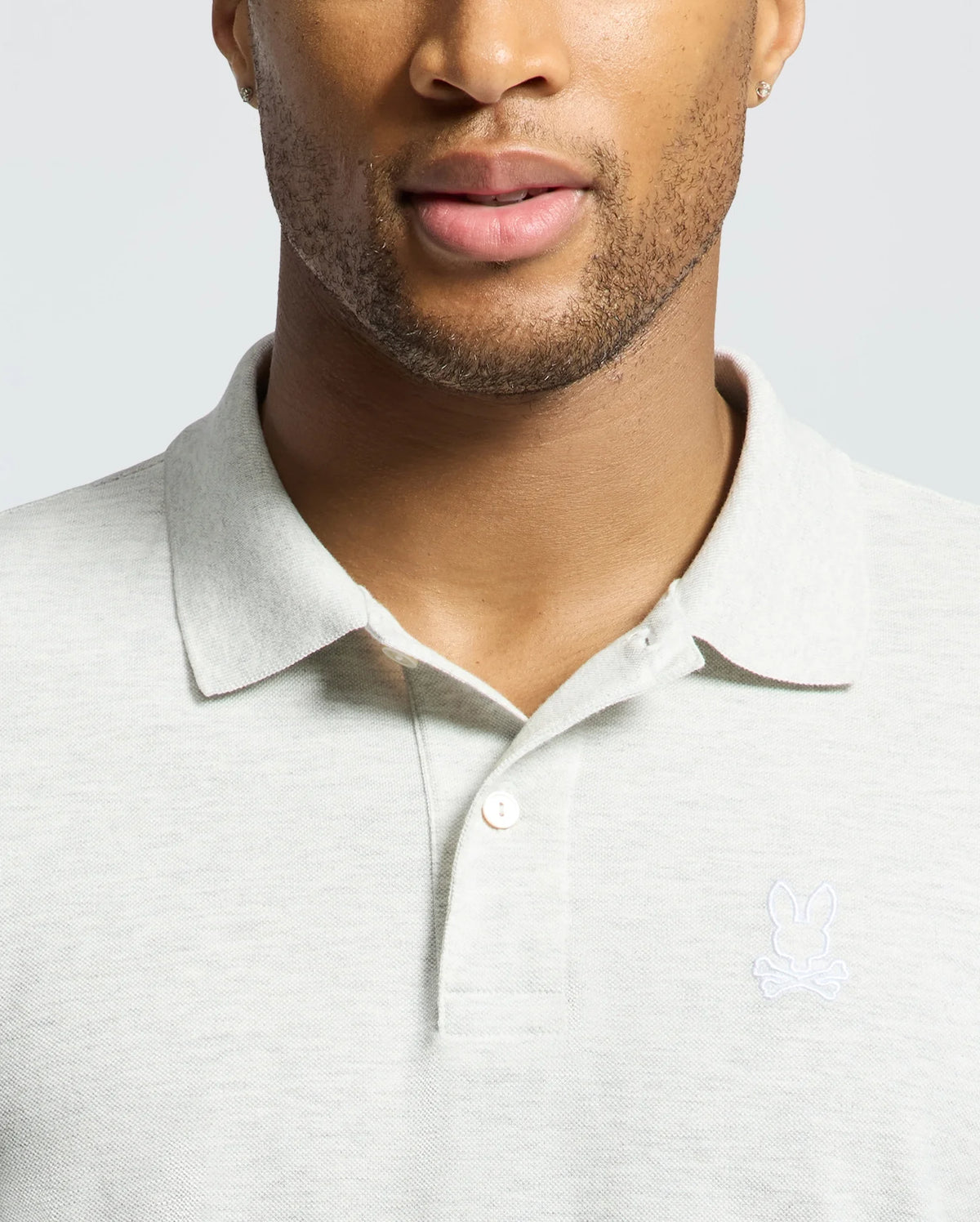 A man wears a Psycho Bunny MENS JAMES PIQUE POLO - B6K945D200 in light gray with an embroidered rabbit and crossed bones logo. His partially visible face shows trimmed facial hair. Made from soft Pima cotton, this shirt offers a durable, slim fit.