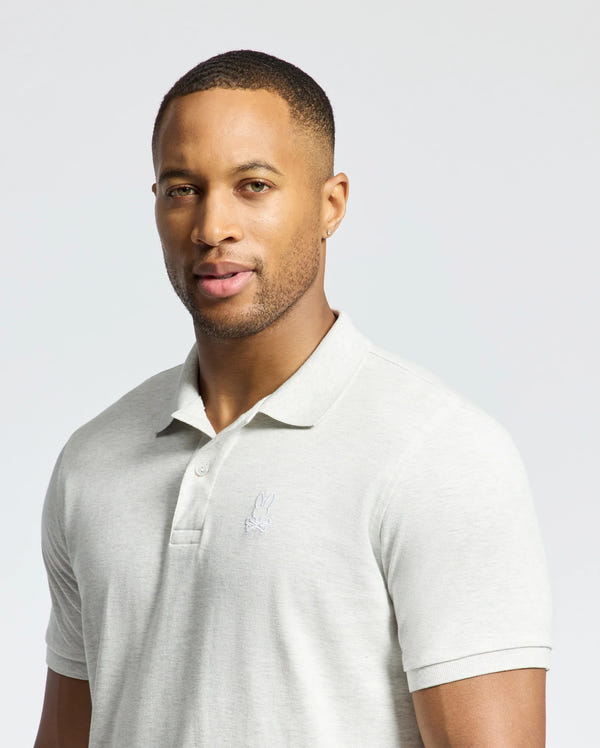 A man in a slim fit Psycho Bunny MENS JAMES PIQUE POLO - B6K945D200, light gray and made from durable Pima cotton, stands against a plain background. The shirt has a small embroidered logo on the left chest. He has short hair and gazes at the camera with a neutral expression.