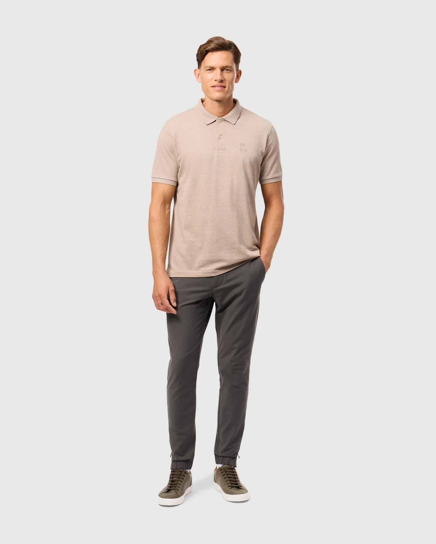 A man stands against a plain white background wearing a Psycho Bunny Men's James Premium Pique Polo Shirt (B6K945D200) made from Pima cotton, dark gray pants, and olive green sneakers. He has one hand in his pocket and is looking towards the camera with a neutral expression.