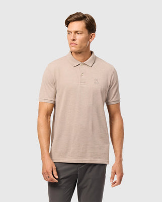A man is standing against a plain background, wearing a light beige Psycho Bunny James Premium Pique Polo Shirt (B6K945D200) made of Pima cotton, paired with dark grey pants. The polo shirt features a small embroidered logo on the left chest. The man has short hair and is gazing off to his left with a neutral expression.