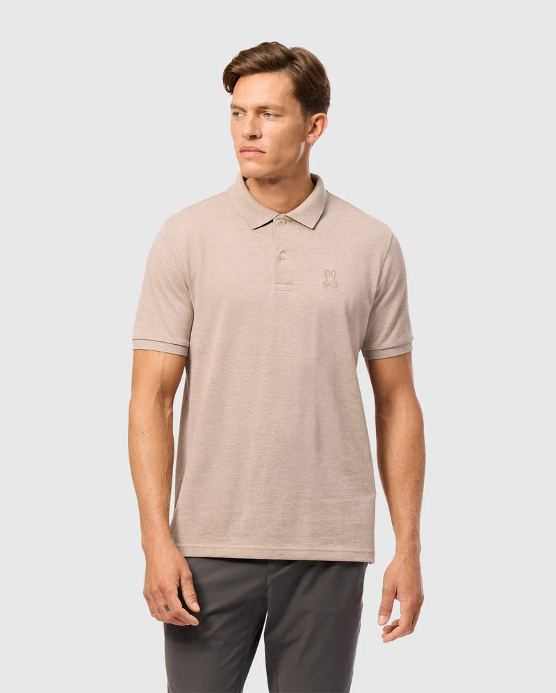 A man is standing against a plain background, wearing a light beige Psycho Bunny James Premium Pique Polo Shirt (B6K945D200) made of Pima cotton, paired with dark grey pants. The polo shirt features a small embroidered logo on the left chest. The man has short hair and is gazing off to his left with a neutral expression.