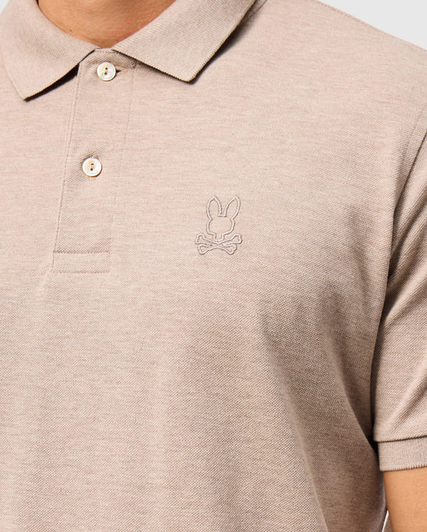 Close-up of a person wearing a beige Psycho Bunny Men's James Premium Pique Polo Shirt (B6K945D200) made from durable Pima cotton, featuring a subtle embroidered bunny head logo with crossbones on the left chest. The shirt has a classic collar and two buttons.