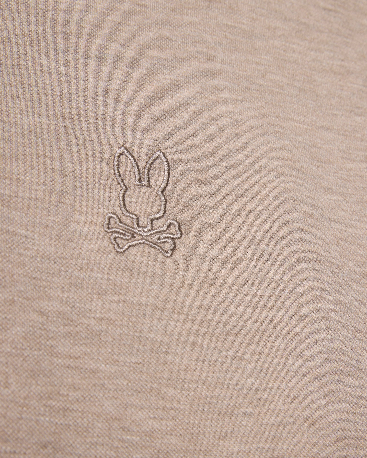 Close-up image of a beige fabric featuring an embroidered logo of a bunny head above two crossed bones, offering a playful twist on the classic skull and crossbones symbol. The embroidery, crafted in a slightly darker shade of beige, enhances the unique character of this durable Psycho Bunny James Premium Pique Polo Shirt - B6K945D200 made from premium Pima cotton.