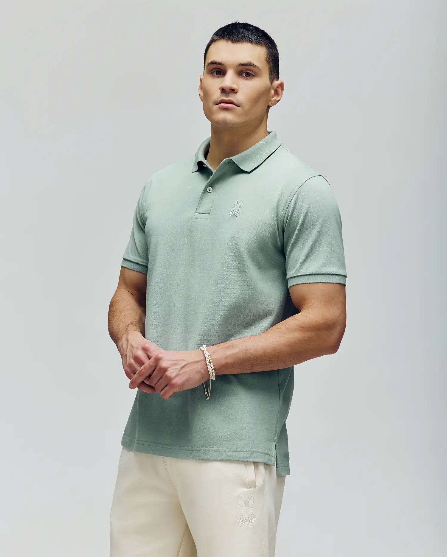 A man with short dark hair and a neutral expression stands against a light background, wearing Psycho Bunny's MENS JAMES PREMIUM PIQUE POLO - B6K945D200 in pale green with a crease-resistant collar and cream-colored pants.
