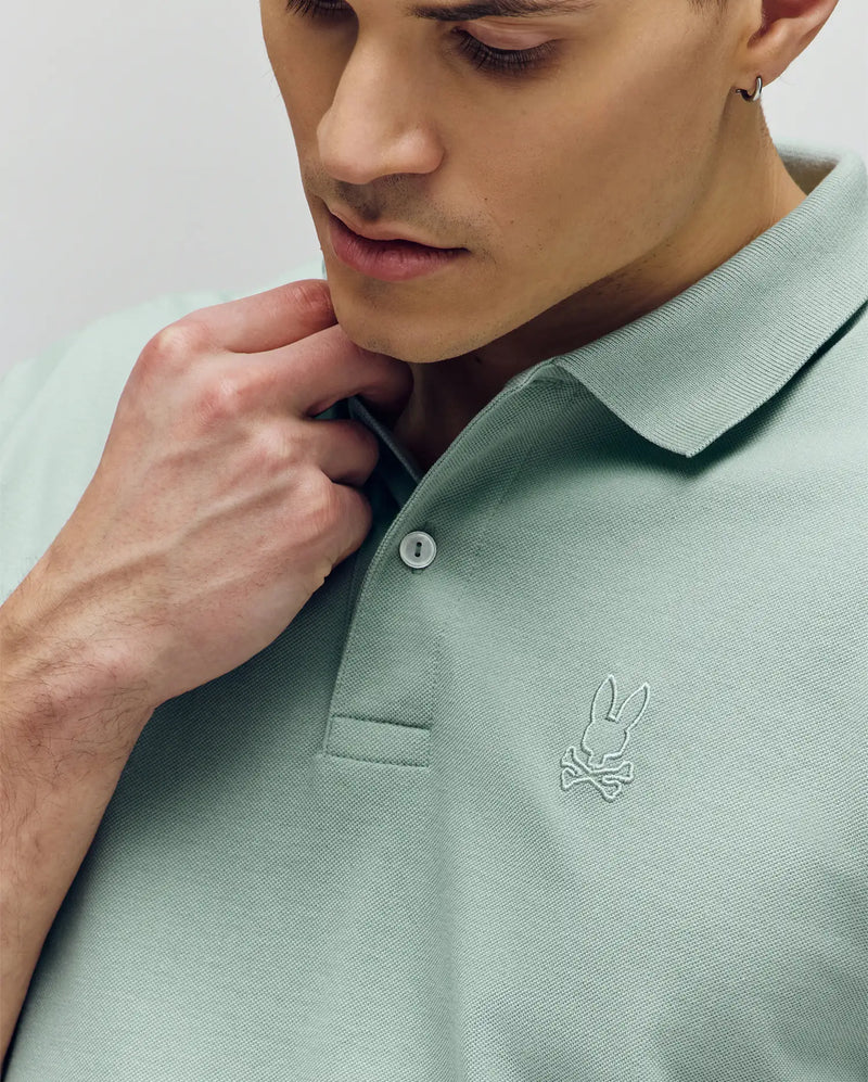 A man wears the Psycho Bunny Mens James Premium Pique Polo (B6K945D200) in light green, featuring an embroidered bunny and crossbones logo on the chest, while looking down and softly holding its crease-resistant collar.