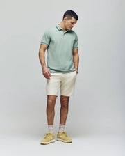 A man stands against a light gray background wearing the Psycho Bunny MENS JAMES PREMIUM PIQUE POLO - B6K945D200 in light green, paired with cream shorts, beige socks, and yellow sneakers. He has one hand in his pocket and looks down to his left.