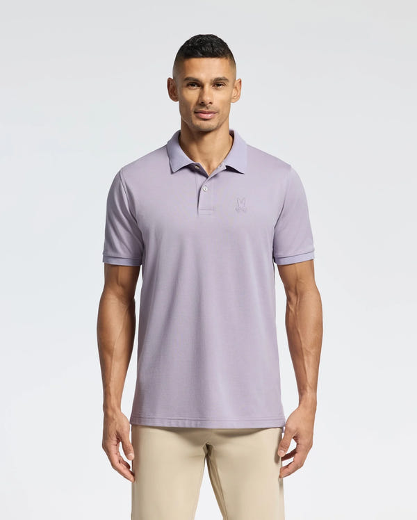 A man stands against a plain background wearing the Psycho Bunny MENS JAMES PIQUE POLO - B6K945D200 in light purple. The slim-fit Pima cotton shirt features a small embroidered logo, and his casual look is complemented by short dark hair and beige pants.