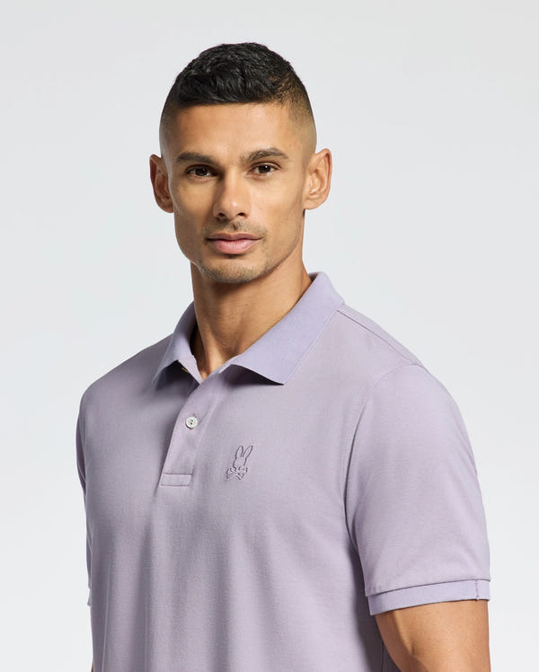 A man stands against a white background wearing a Psycho Bunny MENS JAMES PIQUE POLO - B6K945D200 in light purple, featuring a small embroidered logo on the chest. Made from durable Pima cotton, he has short dark hair and looks at the camera with a neutral expression.