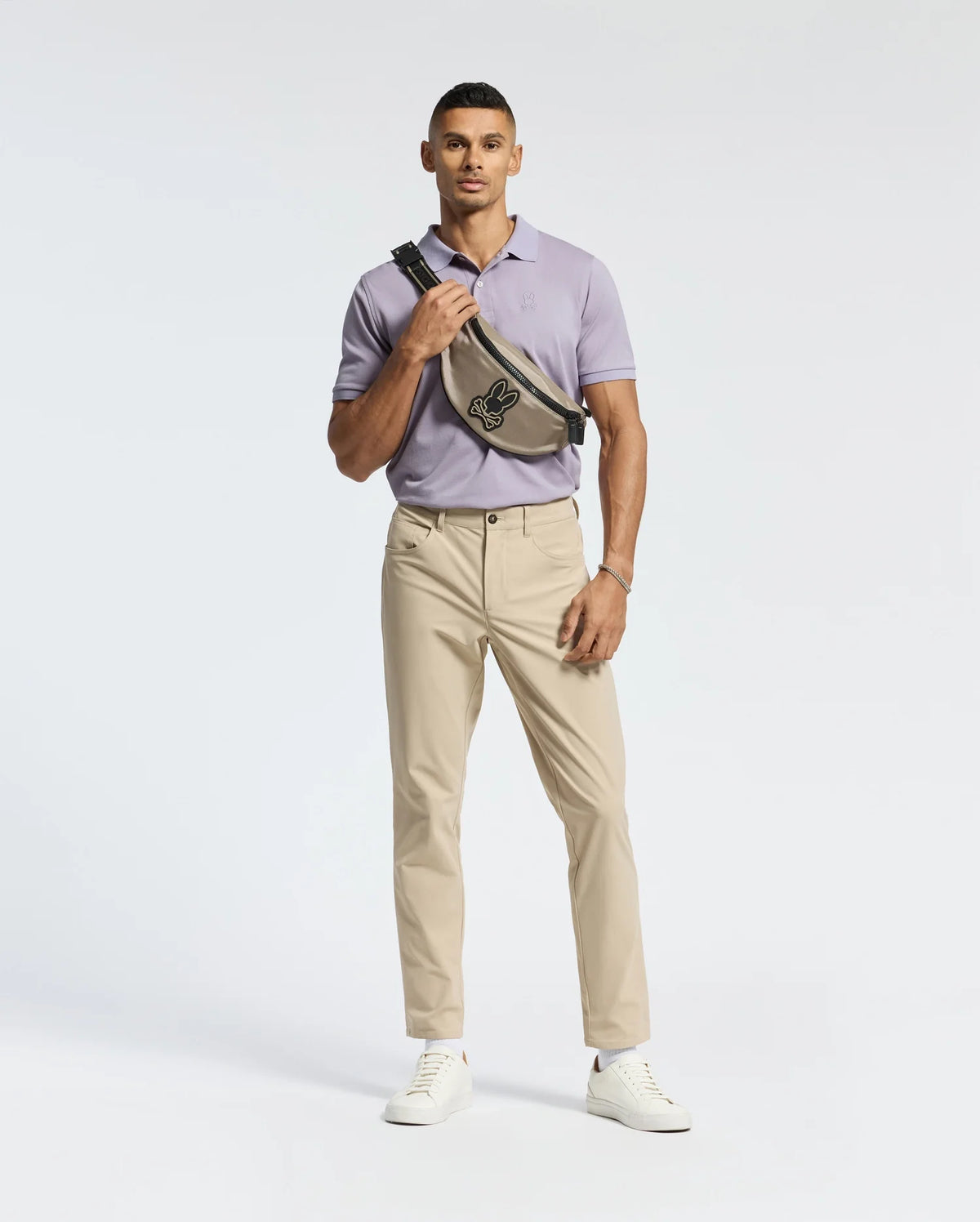 A person confidently wears a slim fit MENS JAMES PIQUE POLO by Psycho Bunny in light purple, paired with beige pants and white sneakers. A black crossbody bag with a bird motif is slung across their chest against a plain white background.