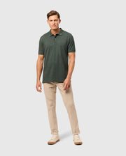 A man stands against a plain background, wearing a dark green Psycho Bunny MENS JAMES PREMIUM PIQUE POLO SHIRT - B6K945D200, beige pants, and light-colored sneakers. He has short hair and is slightly smiling, with his left hand resting by his side and his right hand in his pocket.