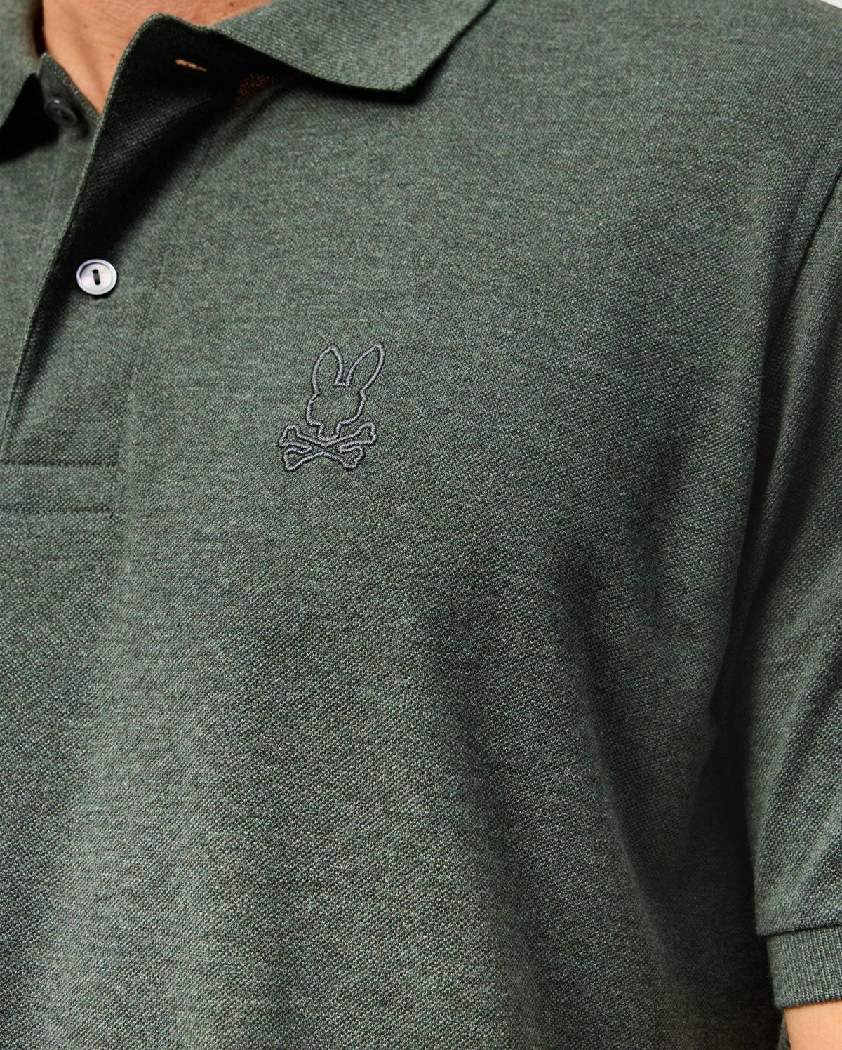 Close-up of a person wearing a dark green Psycho Bunny Men’s James Premium Pique Polo Shirt, model B6K945D200, with two buttons and a small embroidered logo of a rabbit sitting above crossbones on the left chest. The high-quality polo for men features textured fabric, and the image focuses on the upper torso area.
