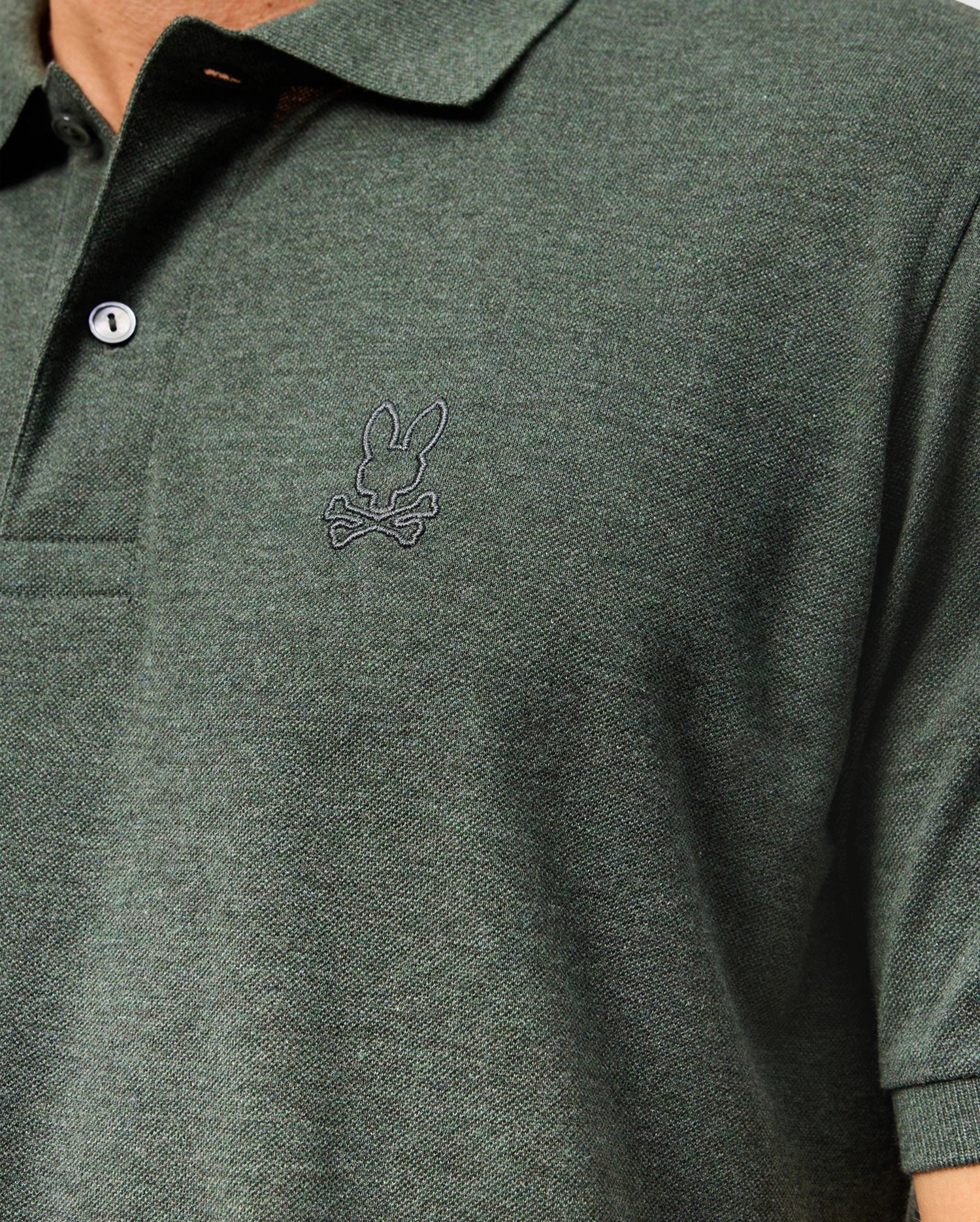 Close-up of a person wearing a dark green Psycho Bunny Men’s James Premium Pique Polo Shirt, model B6K945D200, with two buttons and a small embroidered logo of a rabbit sitting above crossbones on the left chest. The high-quality polo for men features textured fabric, and the image focuses on the upper torso area.
