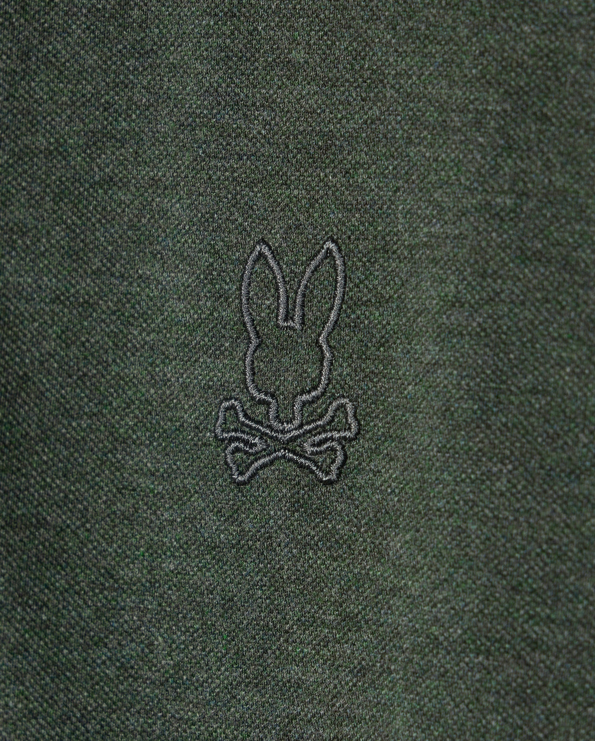 A close-up of the dark green fabric on the Psycho Bunny MENS JAMES PREMIUM PIQUE POLO SHIRT - B6K945D200, showcasing an embroidered black outline of a rabbit head with long ears, positioned above a pair of crossed bones.