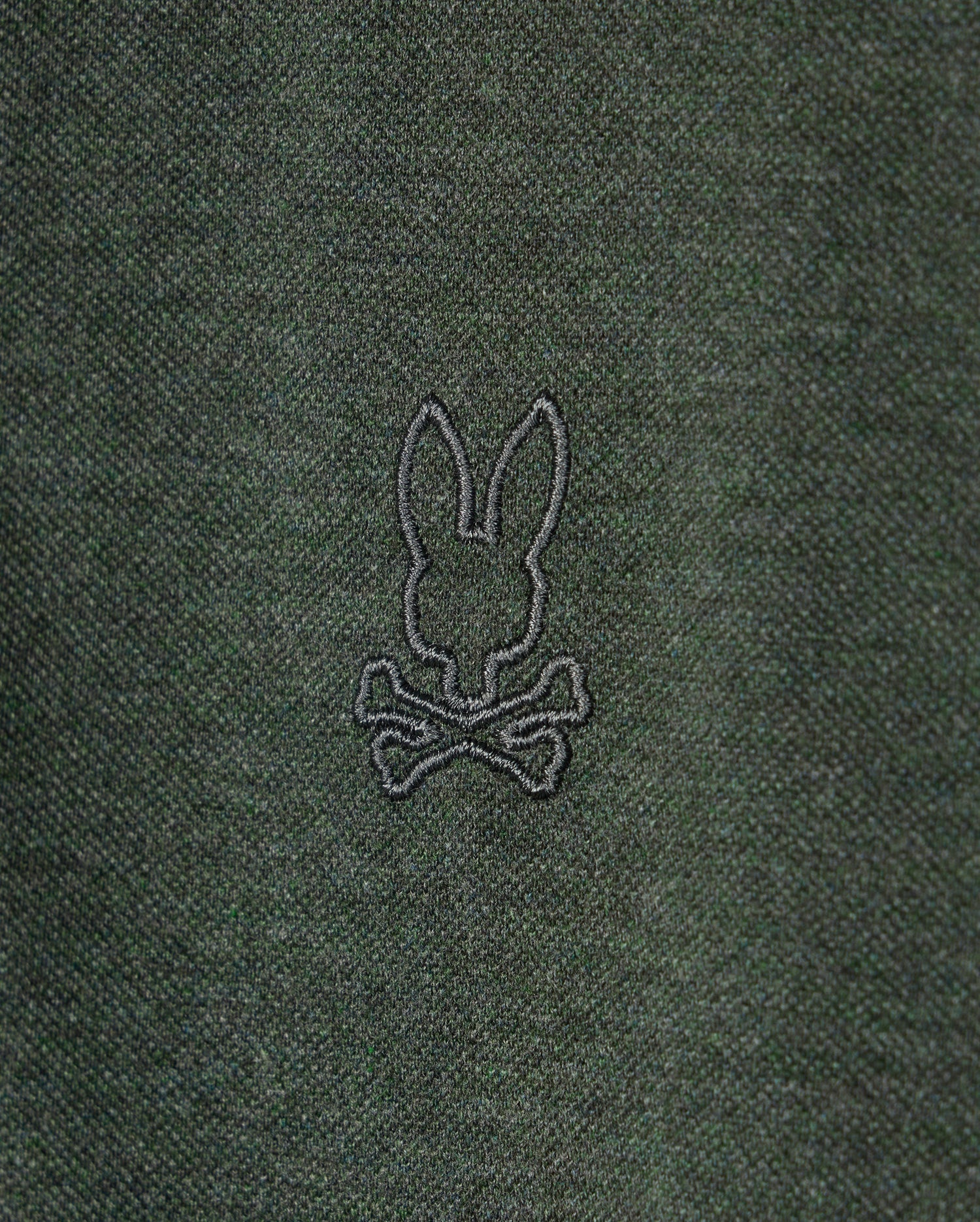 A close-up of the dark green fabric on the Psycho Bunny MENS JAMES PREMIUM PIQUE POLO SHIRT - B6K945D200, showcasing an embroidered black outline of a rabbit head with long ears, positioned above a pair of crossed bones.