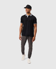 A man is standing against a plain background wearing a Psycho Bunny MENS MALTON PIQUE POLO SHIRT - B6K950D200, featuring black Pima cotton with white trim on the collar and sleeve edges. He pairs it with grey pants, white sneakers, and a black baseball cap. He is looking slightly to the side with his arms relaxed by his sides.