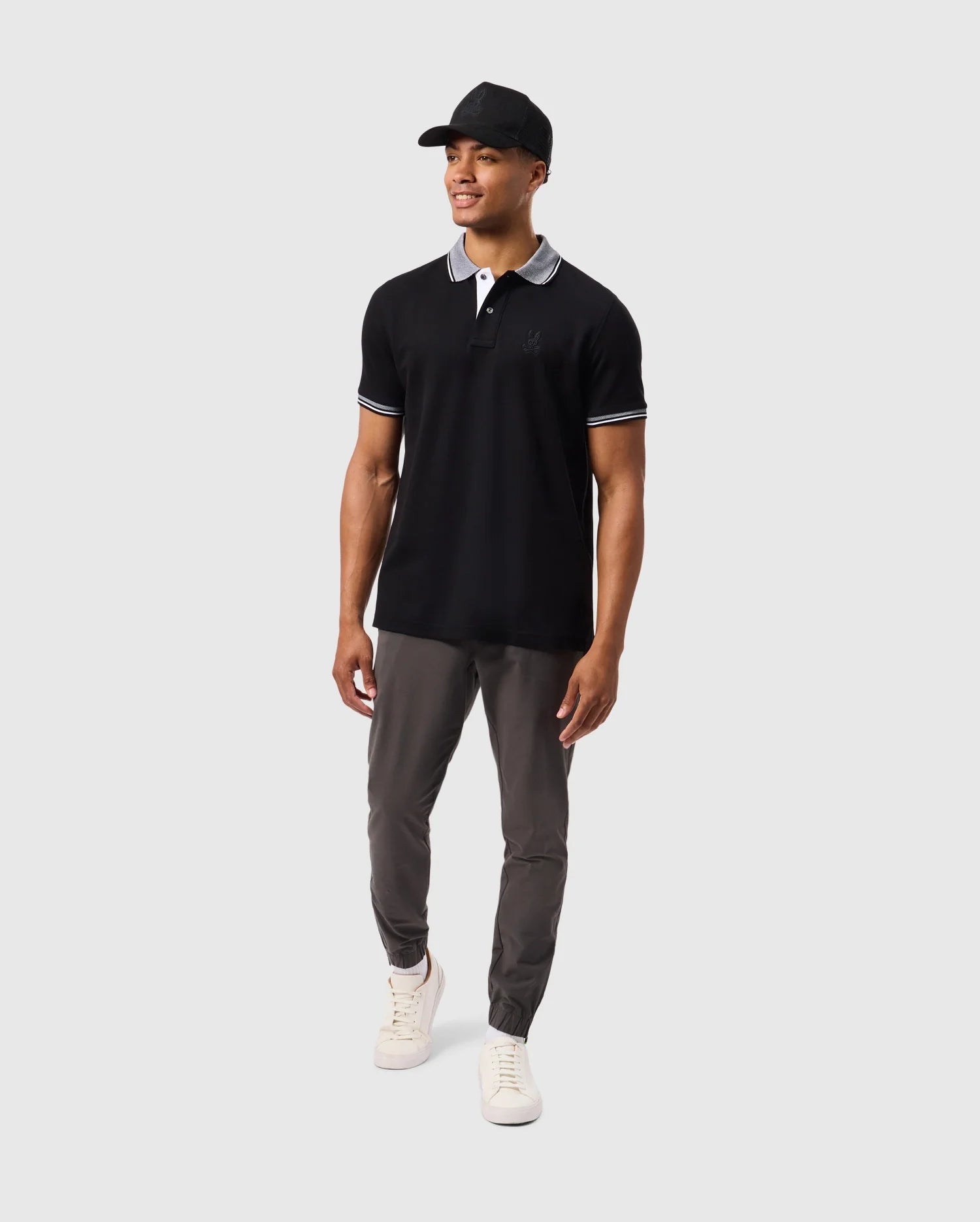 A man is standing against a plain background wearing a Psycho Bunny MENS MALTON PIQUE POLO SHIRT - B6K950D200, featuring black Pima cotton with white trim on the collar and sleeve edges. He pairs it with grey pants, white sneakers, and a black baseball cap. He is looking slightly to the side with his arms relaxed by his sides.