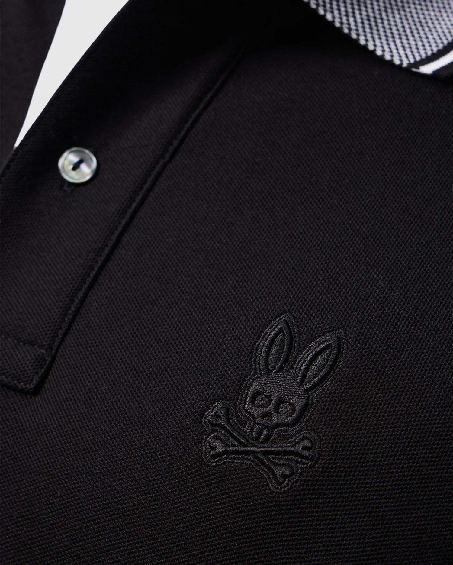 Close-up of the Psycho Bunny Men's Malton Pique Polo Shirt in black Pima cotton, showcasing an embroidered Psycho Bunny skull and crossbones logo on the left chest. The collar features a white and black striped pattern, and the shirt is designed with a button.