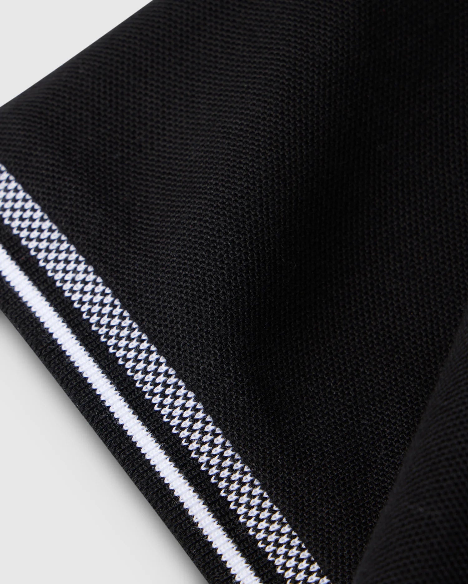 Close-up of a black fabric with an intricate, diamond-knit texture from the Psycho Bunny MENS MALTON PIQUE POLO SHIRT - B6K950D200. The edge showcases a striking white and black patterned border that forms a contrasting stripe. The fabric exudes softness and has a slightly raised surface, embodying the premium quality typical of Psycho Bunny designs. The background is light-colored.