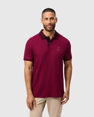 A person wearing a Psycho Bunny Men's Malton Pique Polo Shirt in burgundy, paired with beige pants, stands against a plain background. They have short hair and a beard, embodying classic style with this stylish short sleeve design adorned with an embroidered logo.