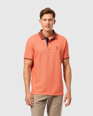 A person wears a Psycho Bunny MENS MALTON PIQUE POLO SHIRT (B6K950D200) with coral color, dark collar accents, and intricate embroidery. They pair it with beige pants and stand against a plain background, looking directly at the camera.