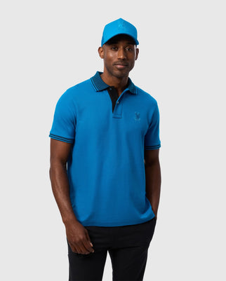 A man is standing against a plain background, wearing a Psycho Bunny MENS MALTON PIQUE POLO SHIRT - B6K950D200 in blue, featuring a black collar and blue trim on the sleeves. He is also donning a matching Psycho Bunny baseball cap. His hands are in his pockets, and he is smiling slightly.