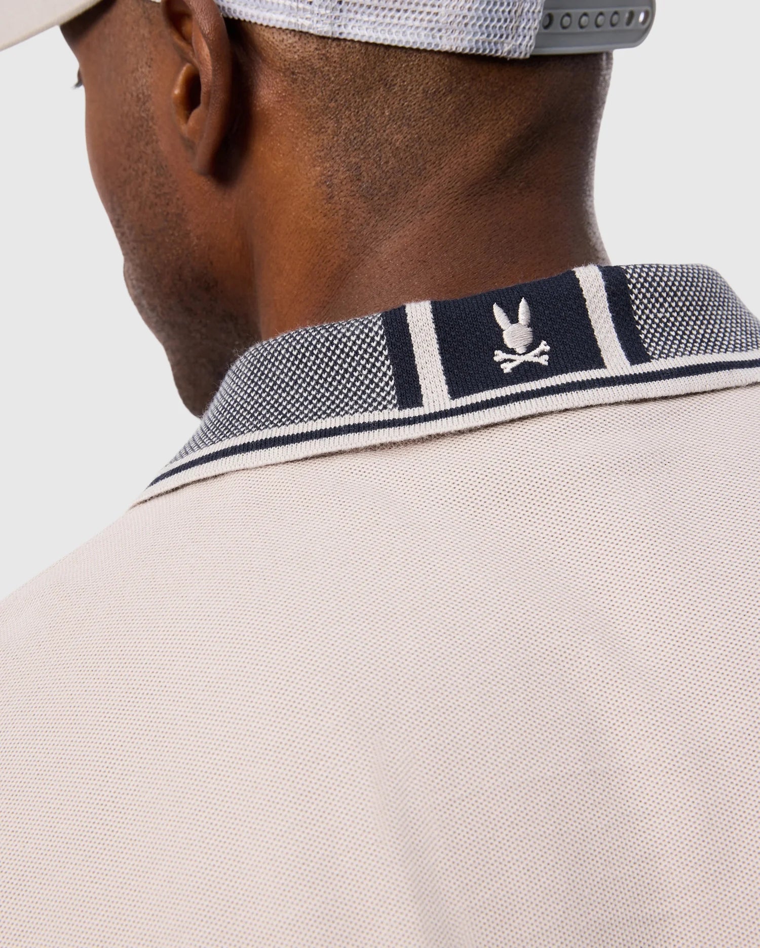 Close-up of the back of a person wearing the light beige MENS MALTON PIQUE POLO SHIRT - B6K950D200 from Psycho Bunny, featuring a navy-collared trim with white stripes and a small Psycho Bunny logo above crossed bones. The shirt is crafted from soft Pima cotton. The person is also wearing a gray cap backward.