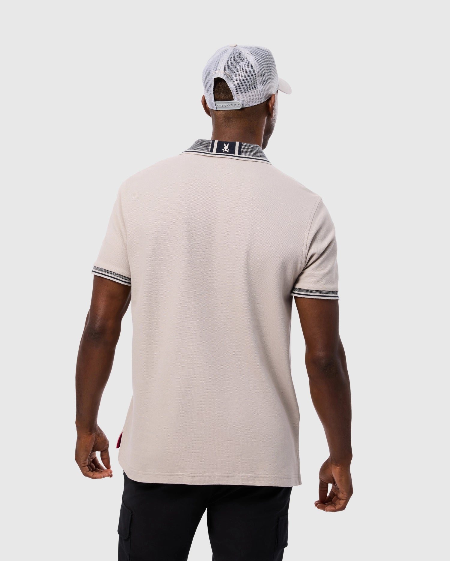 A person is wearing the beige Psycho Bunny MENS MALTON PIQUE POLO SHIRT - B6K950D200, which features black and white detailing on the collar and cuffs, paired with a white baseball cap. The individual, epitomizing the understated elegance of Psycho Bunny, stands facing away from the camera against a plain background.