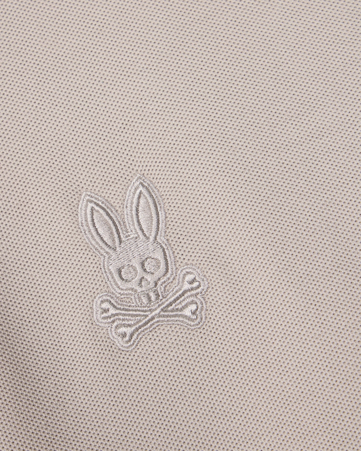 Close-up of the MENS MALTON PIQUE POLO SHIRT - B6K950D200 fabric in a diamond pique pattern, featuring an embroidered skull and crossbones design with rabbit ears, encapsulating the playful Psycho Bunny style. The thread color matches the fabric, creating a subtle and integrated appearance.