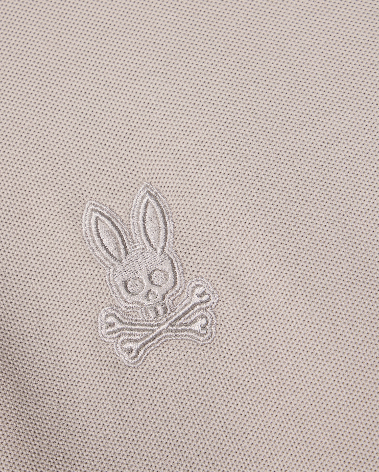 Close-up of the MENS MALTON PIQUE POLO SHIRT - B6K950D200 fabric in a diamond pique pattern, featuring an embroidered skull and crossbones design with rabbit ears, encapsulating the playful Psycho Bunny style. The thread color matches the fabric, creating a subtle and integrated appearance.