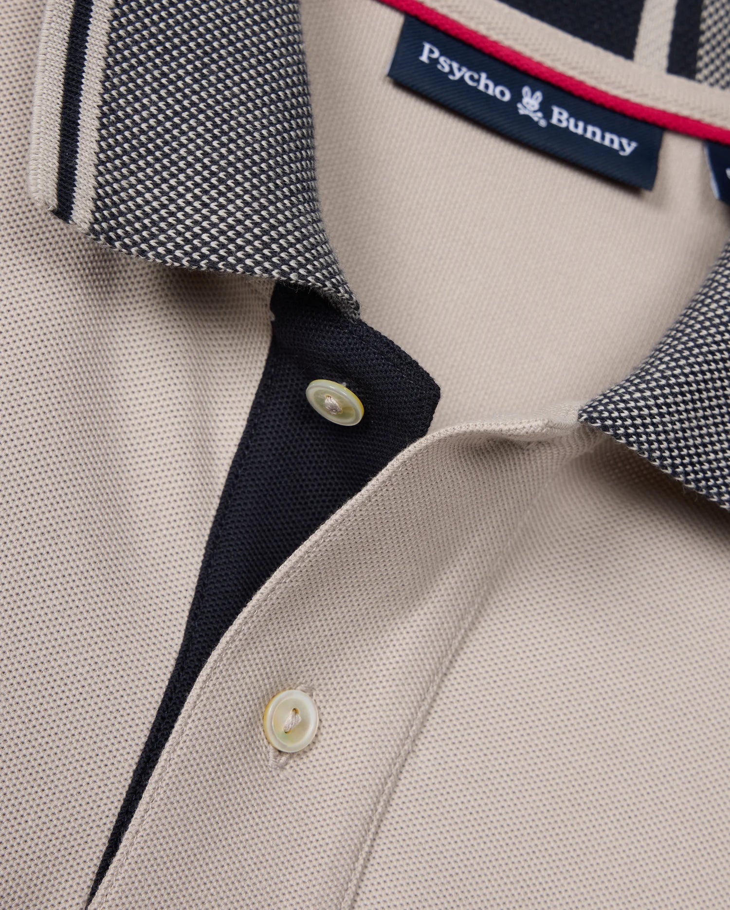 Close-up of a beige Psycho Bunny Malton Pique polo shirt (product code: B6K950D200), featuring a contrasting collar with a black and white checkered pattern. The shirt's placket is partially buttoned with two tan buttons visible, and the collar tag displays the Psycho Bunny logo and brand name.