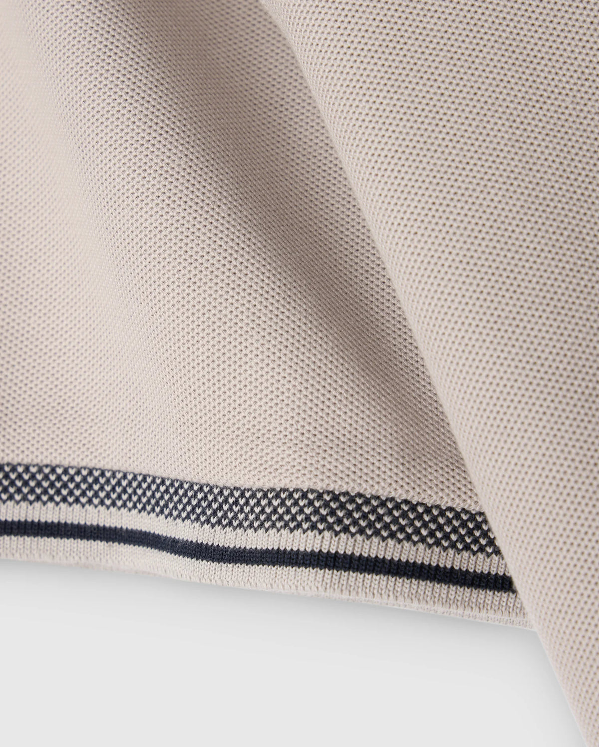 Close-up of the MENS MALTON PIQUE POLO SHIRT - B6K950D200 by Psycho Bunny, featuring a light beige fabric with a textured weave reminiscent of a diamond pique pattern. The edge of the shirt displays a clean, folded hem accented by two thin, parallel black lines running along its length, adding a touch of contrast to the design.