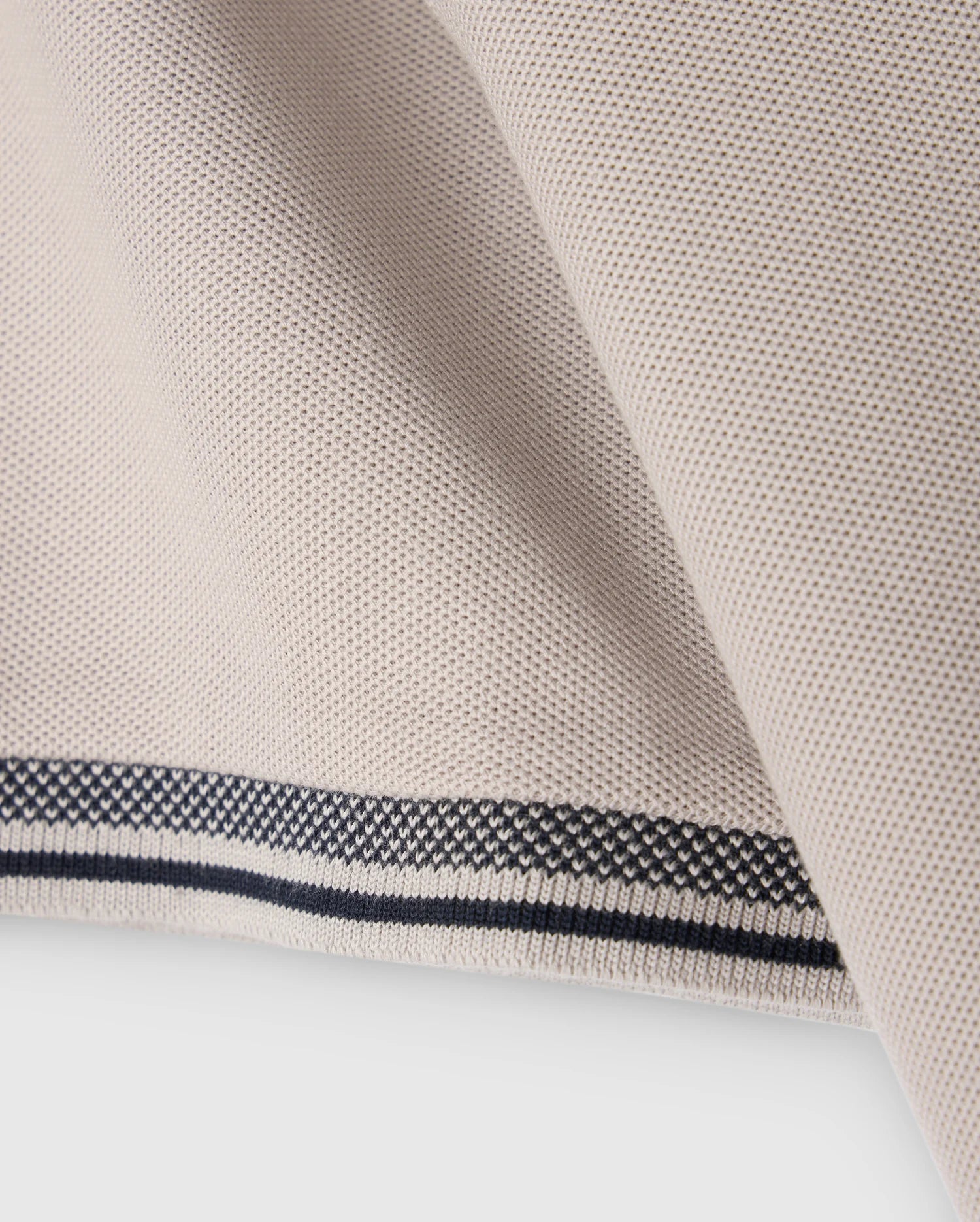 Close-up of the MENS MALTON PIQUE POLO SHIRT - B6K950D200 by Psycho Bunny, featuring a light beige fabric with a textured weave reminiscent of a diamond pique pattern. The edge of the shirt displays a clean, folded hem accented by two thin, parallel black lines running along its length, adding a touch of contrast to the design.