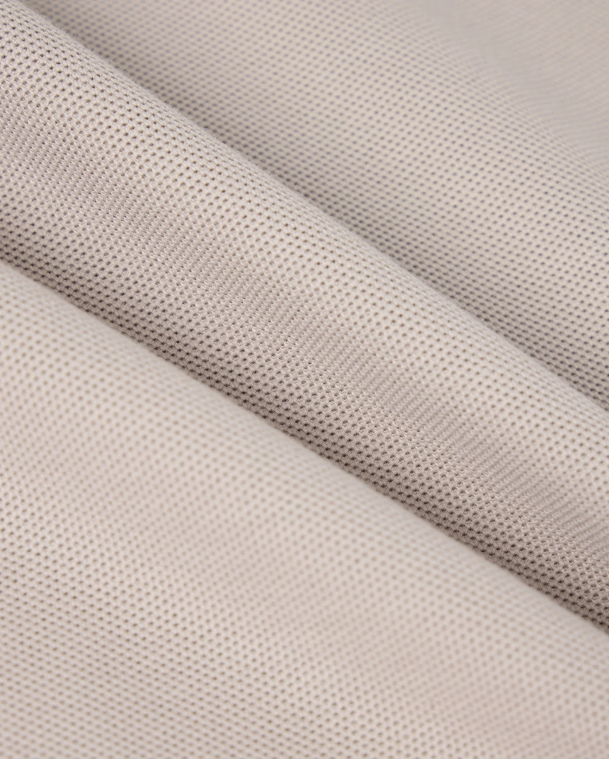 Close-up of a folded piece of beige fabric from the MENS MALTON PIQUE POLO SHIRT - B6K950D200 by Psycho Bunny. The textured material features a fine, even mesh pattern, reminiscent of a diamond pique design. The folds in the fabric create diagonal lines that add depth and dimension to the image. The material appears soft and slightly porous, making it potentially suitable for clothing or upholstery applications.