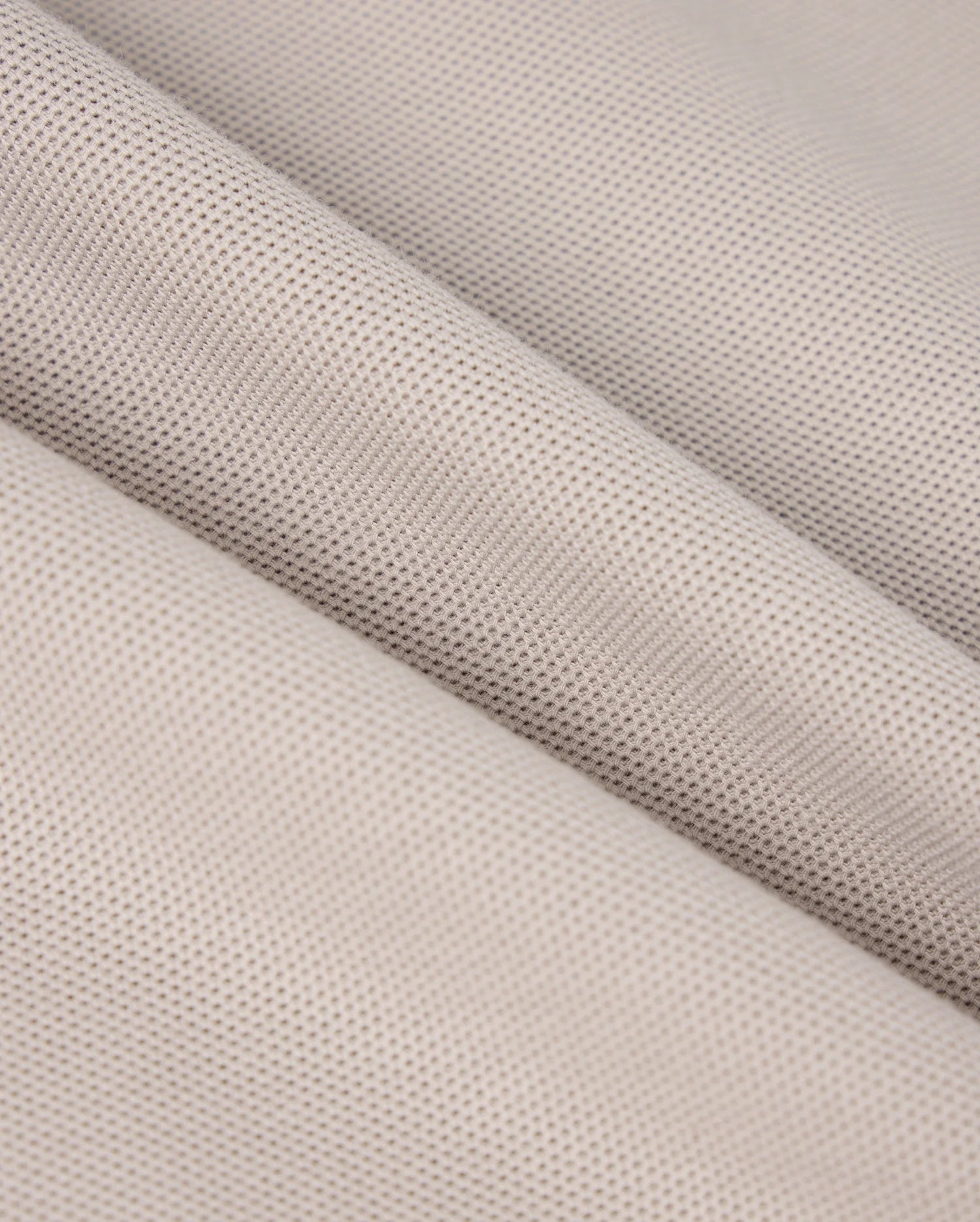 Close-up of a folded piece of beige fabric from the MENS MALTON PIQUE POLO SHIRT - B6K950D200 by Psycho Bunny. The textured material features a fine, even mesh pattern, reminiscent of a diamond pique design. The folds in the fabric create diagonal lines that add depth and dimension to the image. The material appears soft and slightly porous, making it potentially suitable for clothing or upholstery applications.