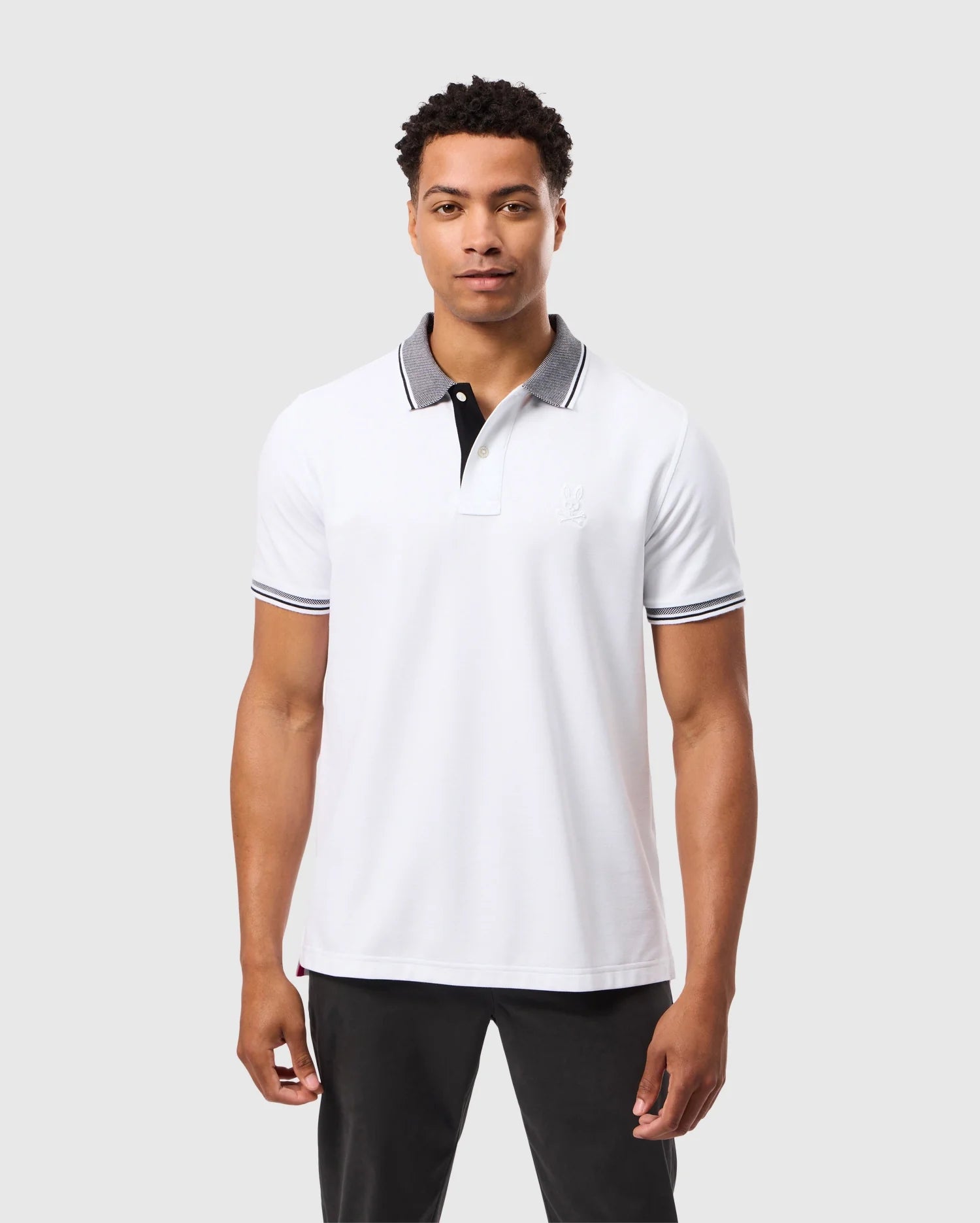 A man dressed in the MENS MALTON PIQUE POLO SHIRT - B6K950D200 by Psycho Bunny, showcasing black accents, stands against a plain background. The polo shirt features a subtle logo on the chest that complements his neutral expression. He pairs it with black pants for a sleek and understated look.
