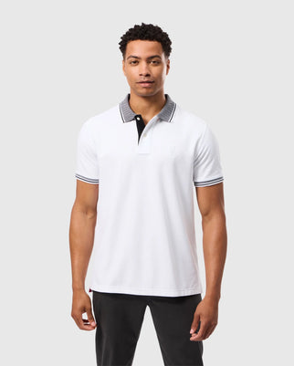 A man dressed in the MENS MALTON PIQUE POLO SHIRT - B6K950D200 by Psycho Bunny, showcasing black accents, stands against a plain background. The polo shirt features a subtle logo on the chest that complements his neutral expression. He pairs it with black pants for a sleek and understated look.