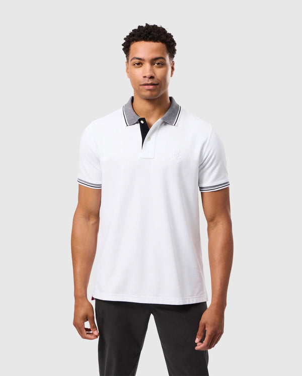 A man dressed in the MENS MALTON PIQUE POLO SHIRT - B6K950D200 by Psycho Bunny, showcasing black accents, stands against a plain background. The polo shirt features a subtle logo on the chest that complements his neutral expression. He pairs it with black pants for a sleek and understated look.