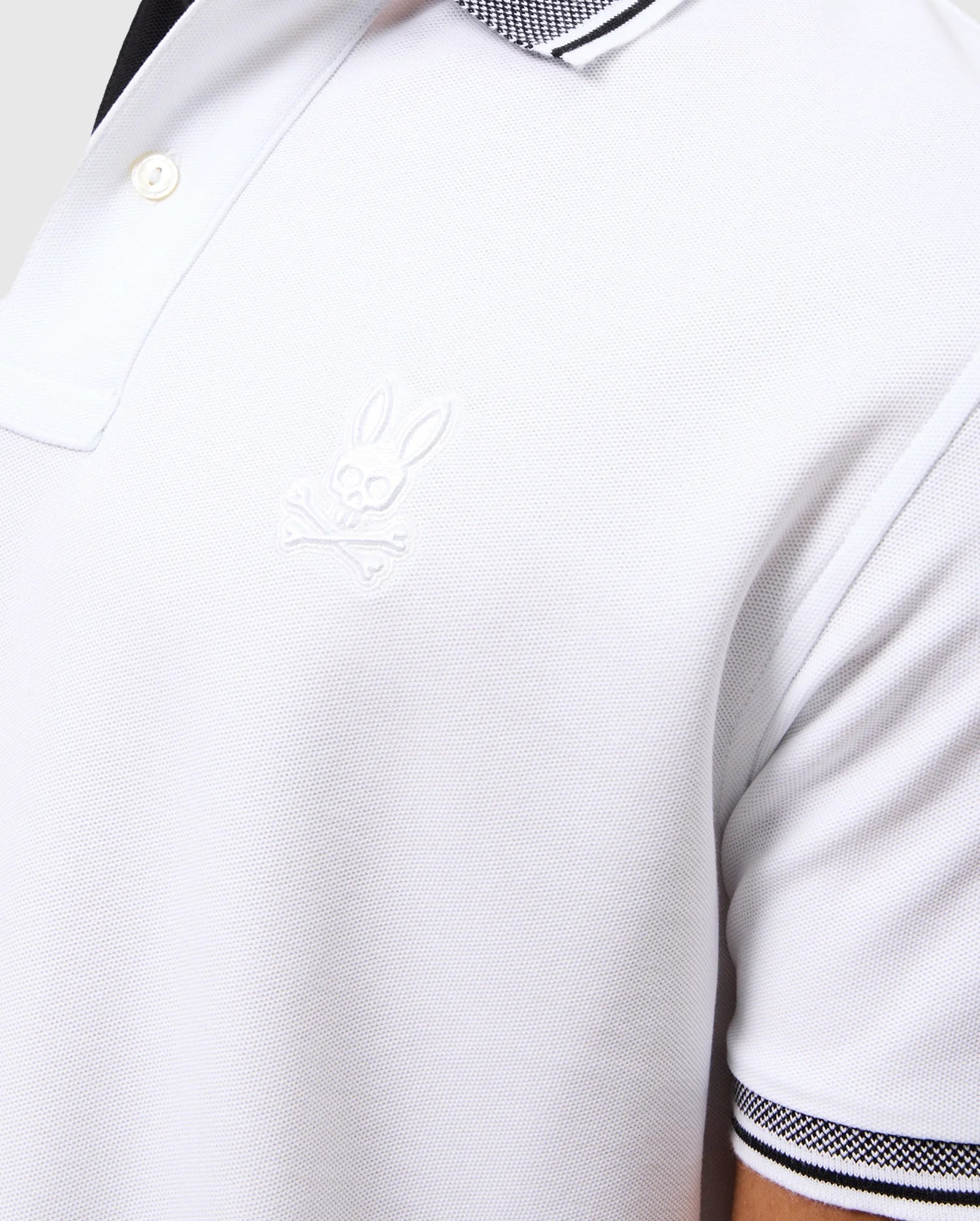 A close-up of a person wearing the MENS MALTON PIQUE POLO SHIRT - B6K950D200 by Psycho Bunny showcases a white Pima cotton polo with black and white striped collar and sleeve accents. The shirt is adorned with an embroidered Psycho Bunny logo depicting a bunny head with crossbones on the chest, all crafted from exquisite fine diamond knit fabric for an added layer of sophistication.