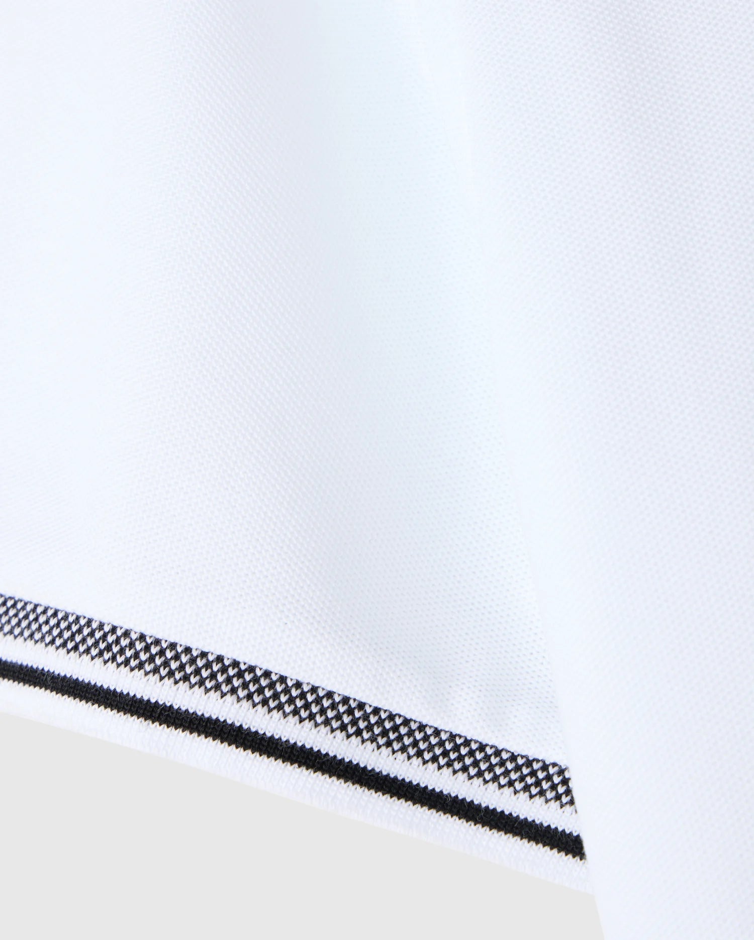 A close-up image of the MENS MALTON PIQUE POLO SHIRT by Psycho Bunny reveals a white fabric with a textured diamond pique pattern, accented by a black and white patterned trim along the edge. The design reflects Psycho Bunny's signature attention to detail, ensuring a smooth and polished finish similar to their timeless Pima cotton polo creations.