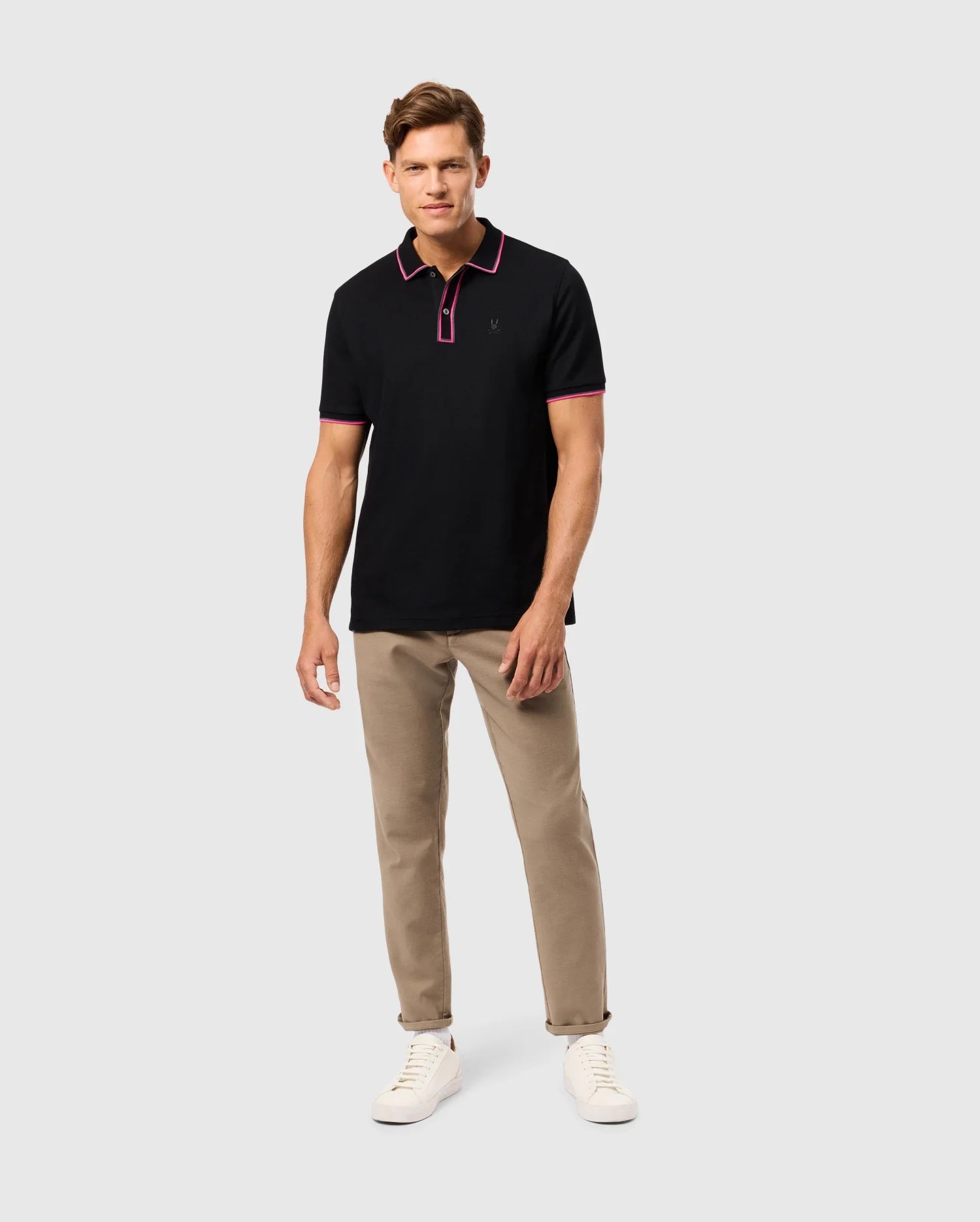 A man stands against a plain background, wearing the MENS KENT PIQUE POLO SHIRT - B6K951D200 by Psycho Bunny, featuring elegant purple trim. He pairs this stylish polo with beige trousers and white sneakers. His hair is styled neatly, complementing the overall polished look as he gazes directly ahead.