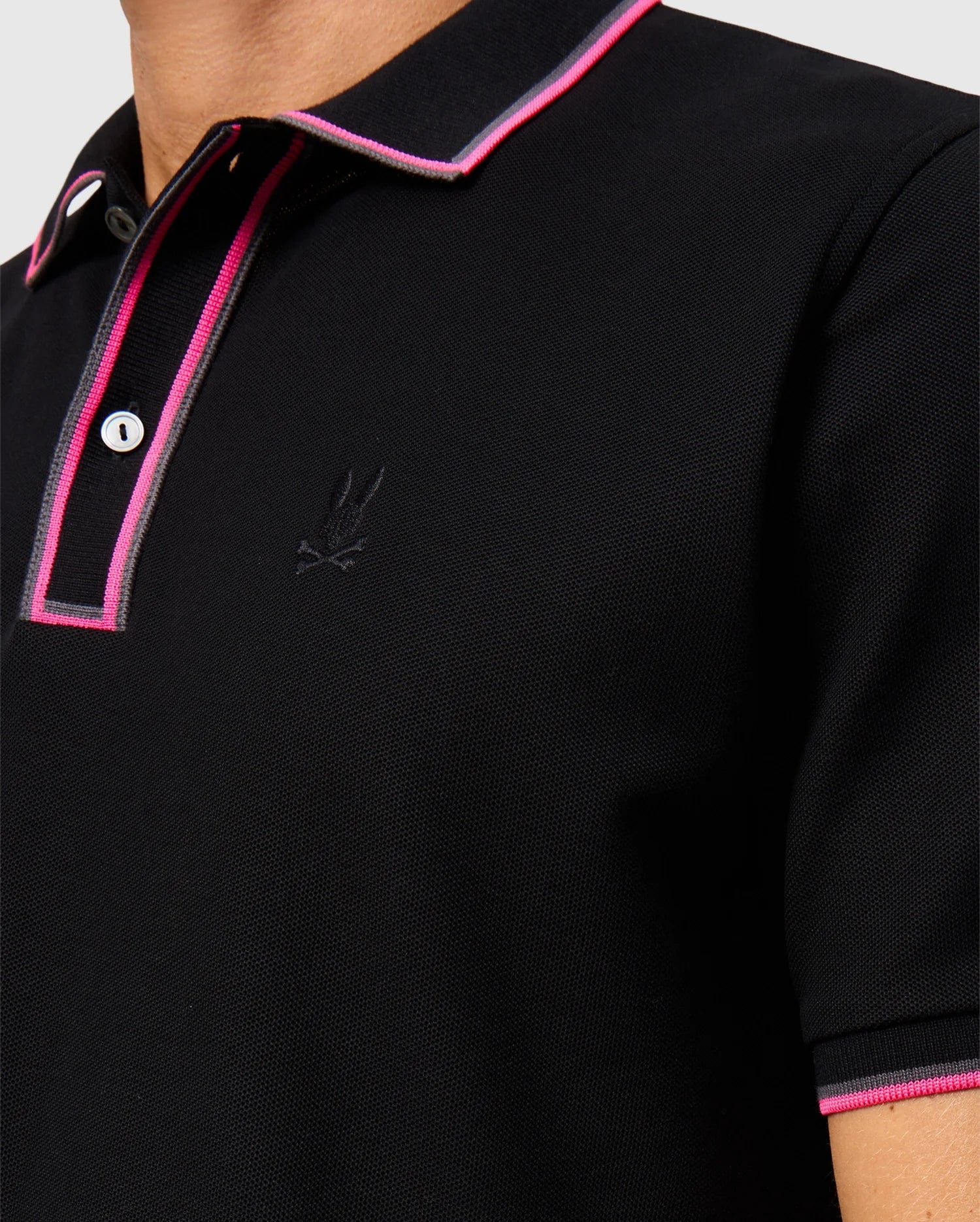 Close-up of a person wearing the Psycho Bunny MENS KENT PIQUE POLO SHIRT - B6K951D200, a stylish black polo shirt with pink trim on the collar and button placket. This pima cotton polo for men features a subtle embroidered logo on the chest, effortlessly blending comfort and fashion.