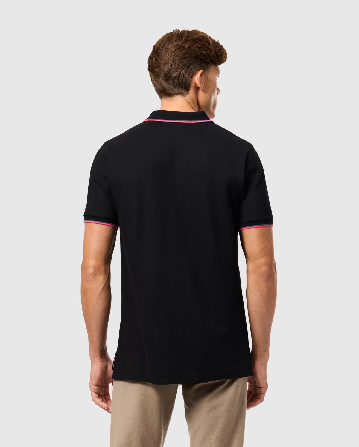 A person is seen from behind wearing the Psycho Bunny MENS KENT PIQUE POLO SHIRT in black, featuring fashionable pink trim on the collar and sleeves. This pima cotton polo shirt for men complements their short brown hair against a simple gray background. Paired with beige pants, it offers a refined yet comfortable style.