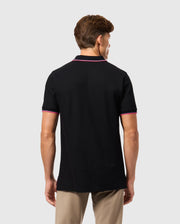 A person is seen from behind wearing the Psycho Bunny MENS KENT PIQUE POLO SHIRT in black, featuring fashionable pink trim on the collar and sleeves. This pima cotton polo shirt for men complements their short brown hair against a simple gray background. Paired with beige pants, it offers a refined yet comfortable style.