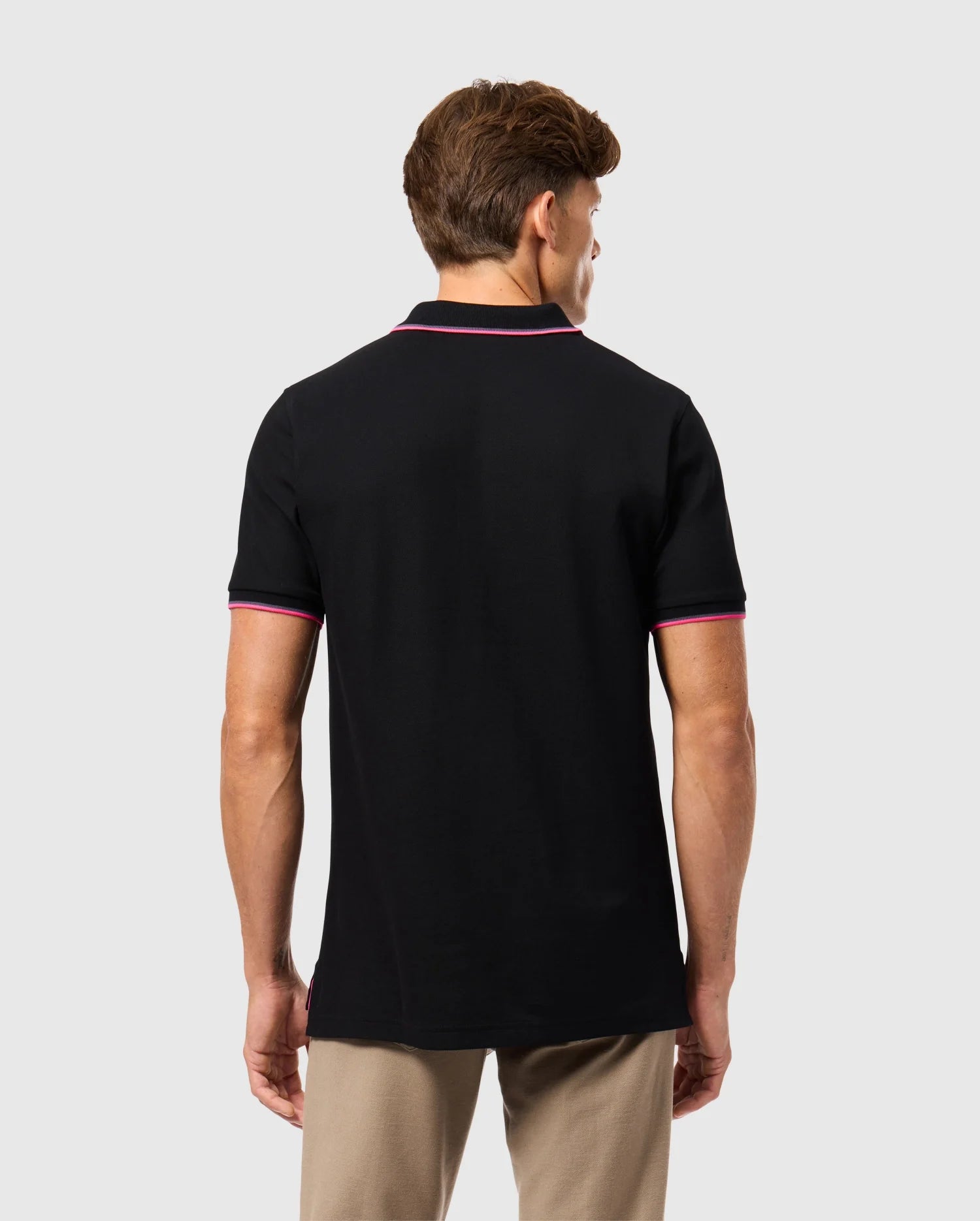 A person is seen from behind wearing the Psycho Bunny MENS KENT PIQUE POLO SHIRT in black, featuring fashionable pink trim on the collar and sleeves. This pima cotton polo shirt for men complements their short brown hair against a simple gray background. Paired with beige pants, it offers a refined yet comfortable style.