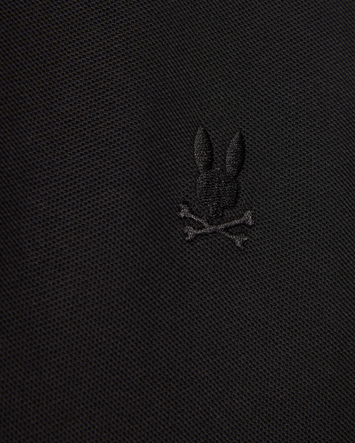 The MENS KENT PIQUE POLO SHIRT - B6K951D200 by Psycho Bunny showcases a close-up view of its fabric, highlighting an embroidered black bunny head above two crossed bones, resulting in a distinctive and edgy motif. The texture of the comfortable pique material is subtly visible.