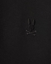 The MENS KENT PIQUE POLO SHIRT - B6K951D200 by Psycho Bunny showcases a close-up view of its fabric, highlighting an embroidered black bunny head above two crossed bones, resulting in a distinctive and edgy motif. The texture of the comfortable pique material is subtly visible.