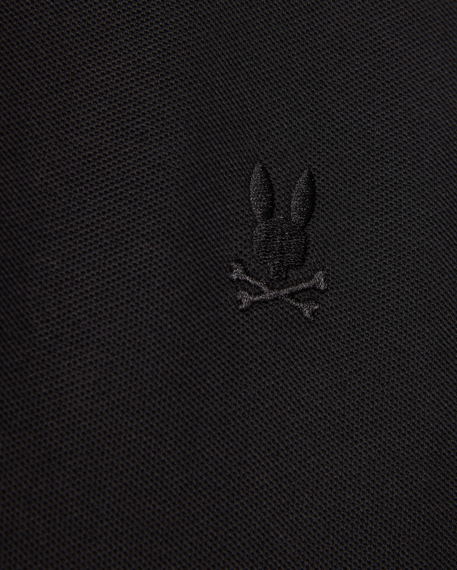 The MENS KENT PIQUE POLO SHIRT - B6K951D200 by Psycho Bunny showcases a close-up view of its fabric, highlighting an embroidered black bunny head above two crossed bones, resulting in a distinctive and edgy motif. The texture of the comfortable pique material is subtly visible.